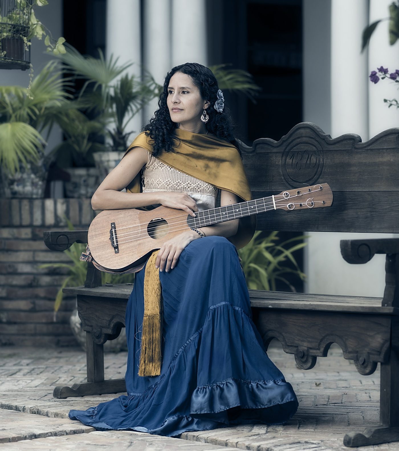 Raquel Palacios Vega seated on a carved wooden bench in a courtyard, her jarana across her lap, wearing a blue skirt and gold rebozo