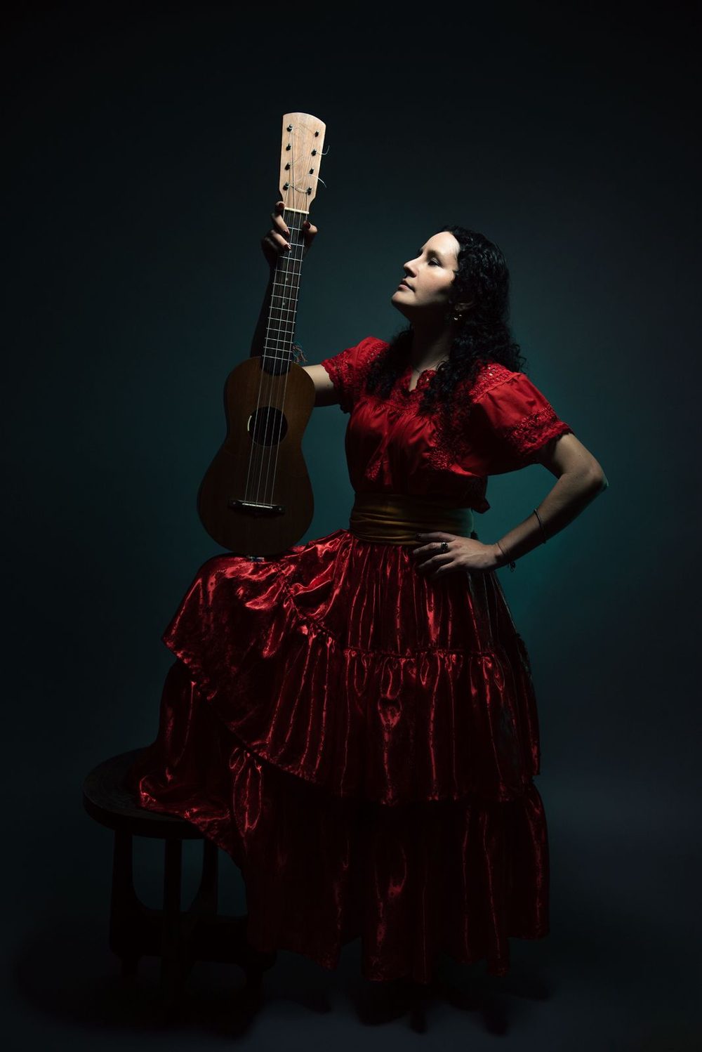 Raquel Palacios Vega in a red traditional dress, holding her jarana aloft against a dark teal background