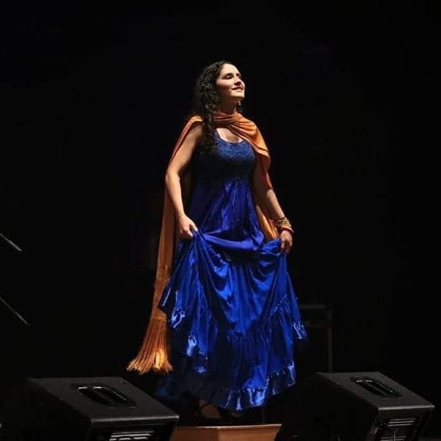Raquel Palacios Vega on stage in a blue skirt and orange rebozo, mid-zapateado